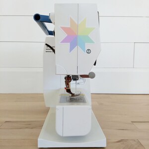 May include: A white sewing machine with a blue handle and a colorful star design on the front. The machine has a needle, presser foot, and a base. The machine is on a wooden floor.