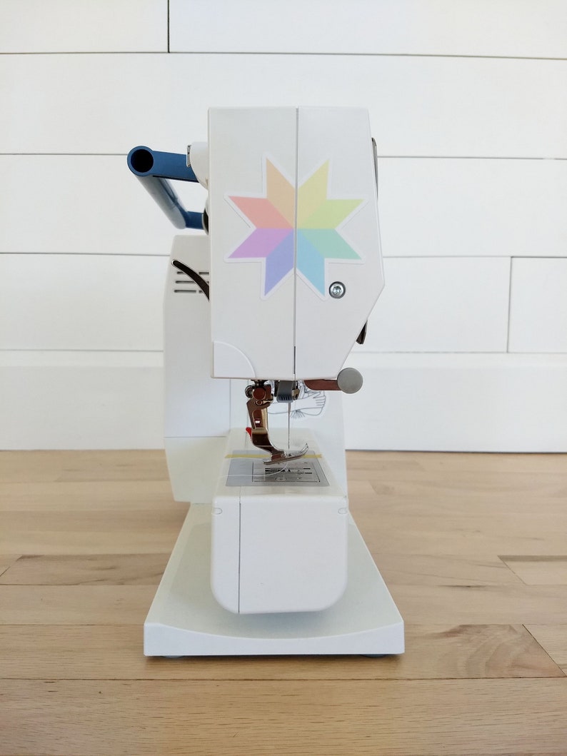 May include: A white sewing machine with a blue handle and a colorful star design on the front. The machine has a needle, presser foot, and a base. The machine is on a wooden floor.