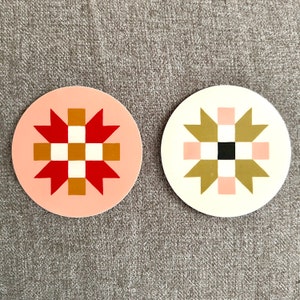 May include: Two round coasters with geometric designs. One coaster has a light pink background with a red and gold starburst pattern. The other coaster has an off-white background with a gold and pink starburst pattern.