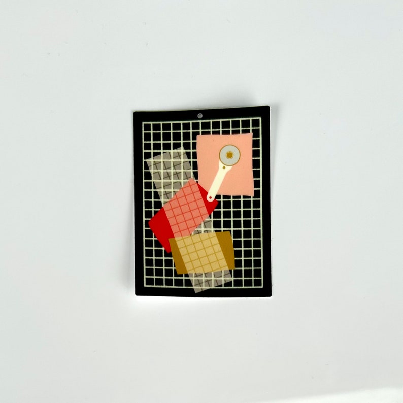 May include: A black rectangular pin with a grid pattern, featuring overlapping squares in red, tan, and transparent materials. A pink square holds a white and silver rotary cutter. The pin is a craft-themed accessory.