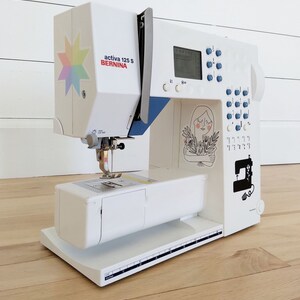 May include: A white Bernina Activa 125 S sewing machine with blue accents and a colorful starburst sticker. The machine features a digital display, control buttons, and decorative illustrations. It sits on a light wood floor.