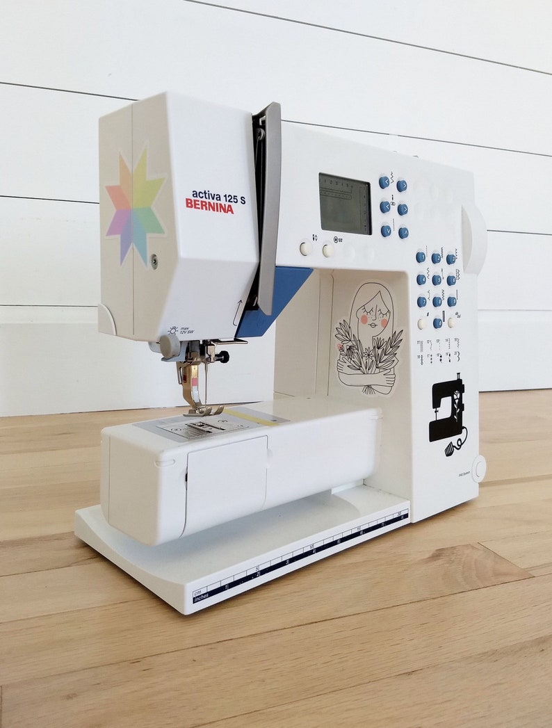 May include: A white Bernina Activa 125 S sewing machine with blue accents and a colorful starburst sticker. The machine features a digital display, control buttons, and decorative illustrations. It sits on a light wood floor.