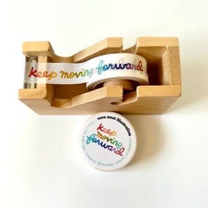 May include: A wooden tape dispenser with a roll of washi tape printed with the words "Keep moving forward" in rainbow hues. A matching sticker with the same design is also present.