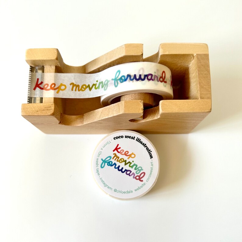 May include: A wooden tape dispenser with a roll of washi tape printed with the words "Keep moving forward" in rainbow hues. A matching sticker with the same design is also present.
