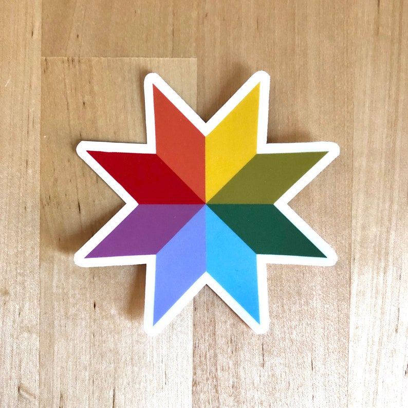 May include: A colorful sticker in the shape of an eight-pointed star. Each point is a different color of the rainbow, including red, orange, yellow, green, blue, and purple. The sticker has a white border and is set against a light wood grain background.