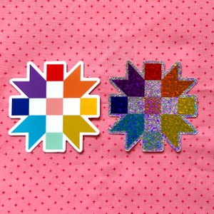 May include: Two colorful star-shaped stickers. One is a rainbow of colors, the other is a glittery version. The stickers are on a pink background with a repeating pattern of small red crosses.