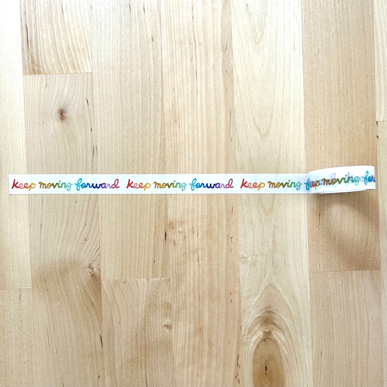 May include: White washi tape printed with the words "keep moving forward" in a rainbow gradient. The tape is unrolled on a light wood surface.