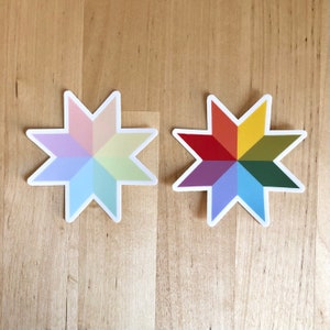 May include: Two star-shaped stickers with a white border. One sticker features pastel colors, while the other displays a vibrant spectrum of colors. The stickers are placed on a light brown wooden surface.