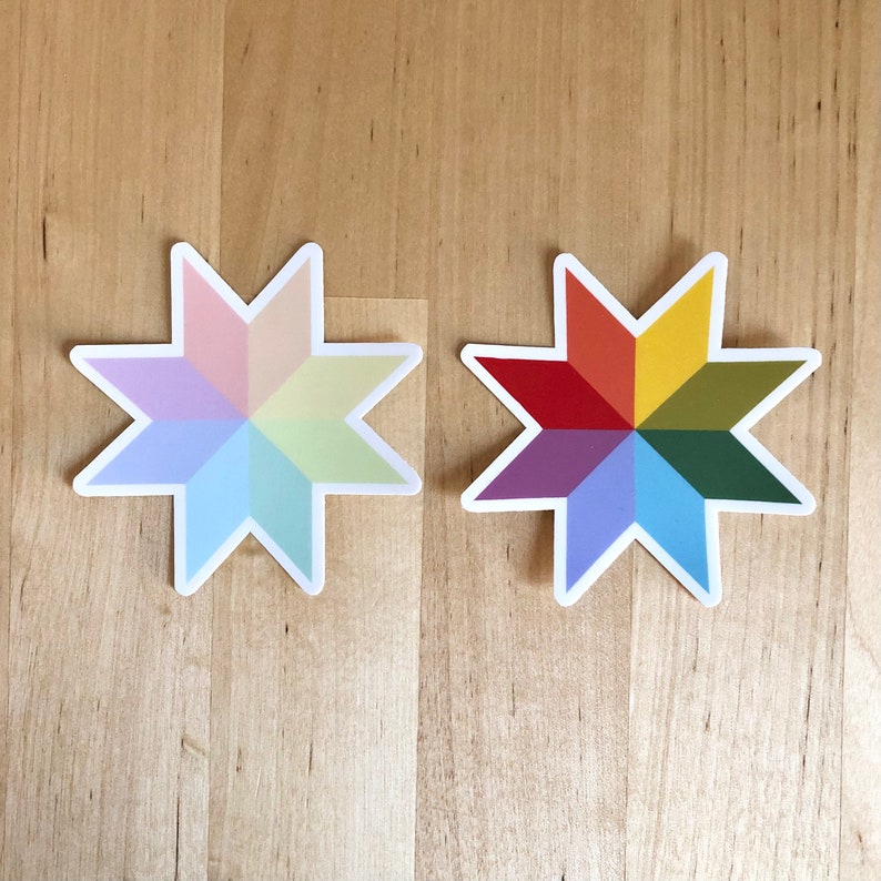 May include: Two star-shaped stickers with a white border. One sticker features pastel colors, while the other displays a vibrant spectrum of colors. The stickers are placed on a light brown wooden surface.