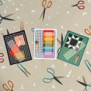 May include: Three colorful cutting mats with rotary cutters, featuring a black, white, and green grid design. The mats display geometric patterns and a rainbow color gradient. The background is a beige fabric with scissors.