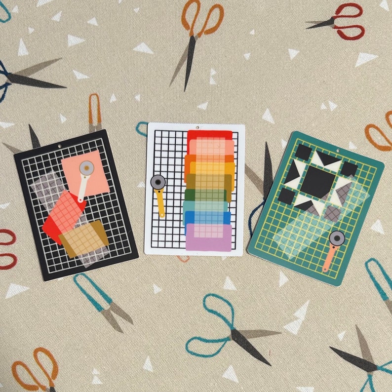 May include: Three colorful cutting mats with rotary cutters, featuring a black, white, and green grid design. The mats display geometric patterns and a rainbow color gradient. The background is a beige fabric with scissors.