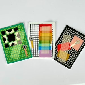 May include: Three colorful quilting-themed cutting mats with rotary cutters. One green mat features a geometric design, the second is white with a rainbow of colors, and the third is black with pink and tan squares. Each mat has a hole for hanging.