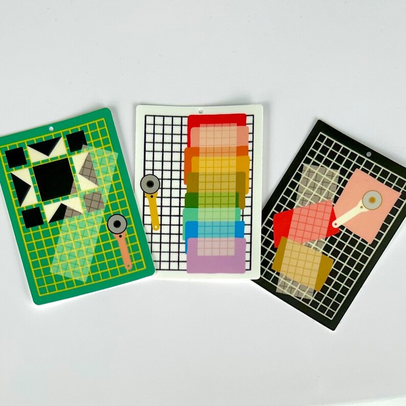 May include: Three colorful quilting-themed cutting mats with rotary cutters. One green mat features a geometric design, the second is white with a rainbow of colors, and the third is black with pink and tan squares. Each mat has a hole for hanging.