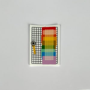 May include: A white rectangular charm with a black grid pattern, a rainbow gradient, and a yellow rotary cutter. The rainbow gradient includes red, orange, yellow, green, blue, and purple. The charm has a small hole at the top.