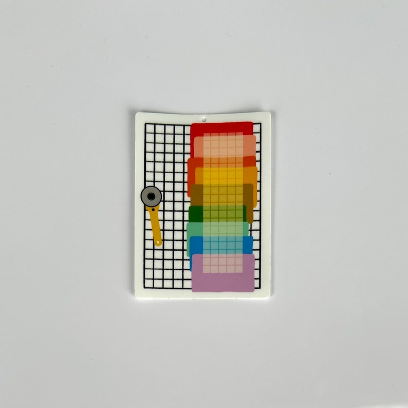 May include: A white rectangular charm with a black grid pattern, a rainbow gradient, and a yellow rotary cutter. The rainbow gradient includes red, orange, yellow, green, blue, and purple. The charm has a small hole at the top.