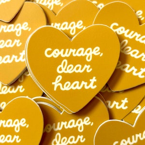 May include: Gold heart-shaped stickers with the phrase "courage, dear heart" in white script. The stickers are scattered, with one in focus. The background is filled with similar stickers, creating a layered effect.