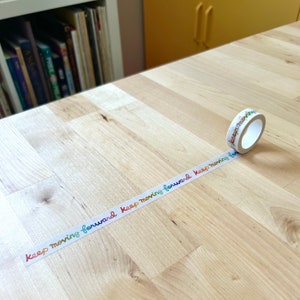 May include: A roll of decorative washi tape with the words "keep moving forward" in a rainbow gradient. The tape is unrolled on a light wood surface.
