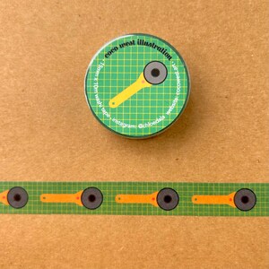 May include: A green and yellow button and washi tape featuring a rotary cutter design. The button has the text "coco west illustration" and social media information. The washi tape has a repeating pattern of the same rotary cutter design.