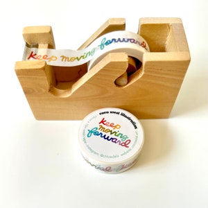 May include: A wooden tape dispenser holding a roll of decorative tape. The tape features the words "Keep Moving Forward" in a rainbow gradient. A separate roll of tape with the same design is also present.