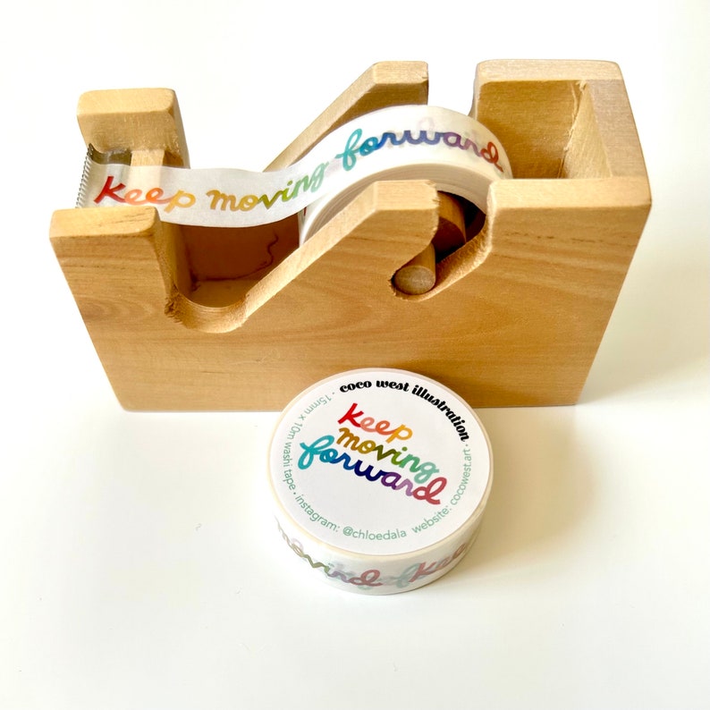 May include: A wooden tape dispenser holding a roll of decorative tape. The tape features the words "Keep Moving Forward" in a rainbow gradient. A separate roll of tape with the same design is also present.