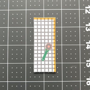 May include: A small, rectangular quilting ruler with a grid pattern and yellow edges. A green rotary cutter with a red and brown blade is positioned on the ruler, suggesting a crafting or sewing project. The ruler is on a black surface.