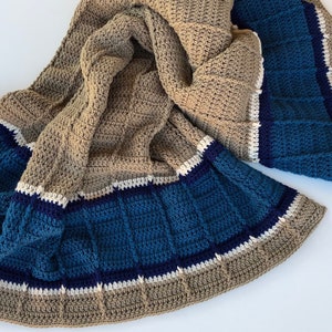 May include: A handmade crochet blanket in shades of tan and blue. The blanket features a textured stitch pattern with a border of dark blue, white, and tan. The blanket is folded and draped, showcasing its design and construction.