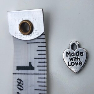 May include: Silver heart-shaped charm with the text "Made with Love" on it. The charm is about 1 inch in size and is shown next to a measuring tape.