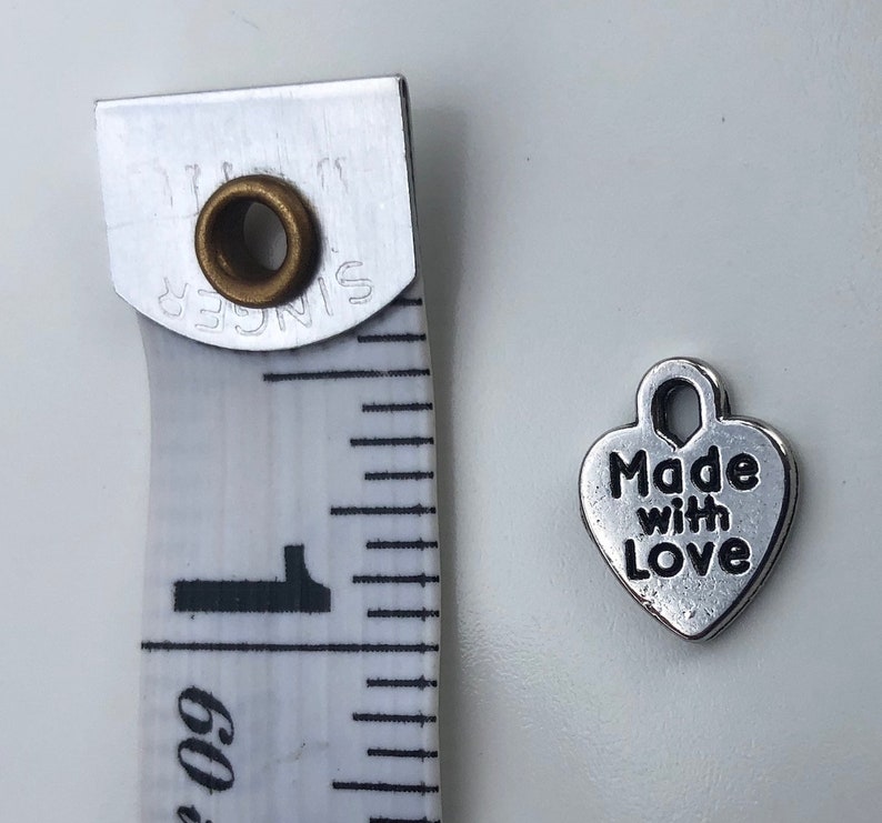 May include: Silver heart-shaped charm with the text "Made with Love" on it. The charm is about 1 inch in size and is shown next to a measuring tape.