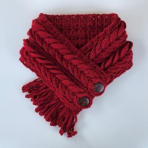 May include: A red crocheted cowl with a cable knit pattern and two wooden buttons. The cowl has a fringe at the bottom.
