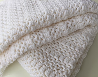 May include: A white crocheted blanket with a textured pattern. The blanket is folded in thirds and is made of soft yarn.