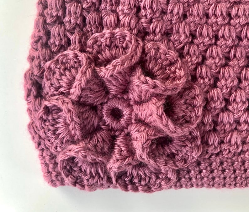 May include: Close-up of a crocheted flower design in a deep pink color. The flower is made up of multiple layers of crocheted stitches, creating a textured and dimensional look.