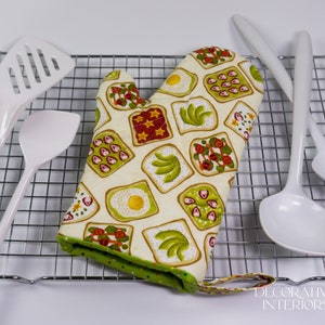 May include: An oven mitt with a whimsical toast design featuring avocado, strawberries, and fried eggs on a cream background. The mitt is surrounded by white kitchen utensils and rests on a metal cooling rack.