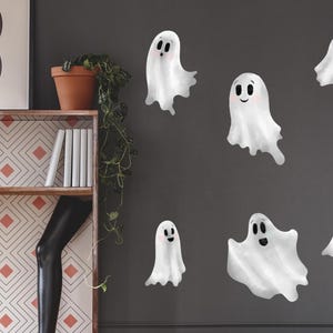 May include: A dark gray wall decorated with white ghost decals. A white sofa with orange and pink pillows sits below the decals. Two small orange tables and a black table are in front of the sofa. A black mannequin leg is visible on the left.