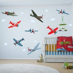 May include: A nursery room with a white crib and a white wall with multiple airplane wall decals. The decals are in various colors, including red, blue, green, and brown. The decals are of different sizes and shapes, and some have clouds around them. There is a teddy bear sitting on the floor in front of the crib.