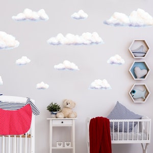 May include: A nursery scene with white cribs, a small white table, and cloud wall decals. The cribs are decorated with red and blue accents. A teddy bear and a plant sit on the table. Decorative shelves with nautical themes are on the wall.