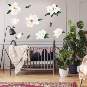 May include: White magnolia flower wall decals with green leaves on a white wall. The decals are arranged in a random pattern. A crib with a white blanket and a pink and white bunting is in the foreground. There are two potted plants in the background.
