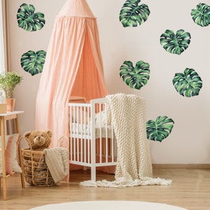May include: A white crib with a pink canopy and a white knitted blanket. The wall is decorated with green monstera leaf decals.