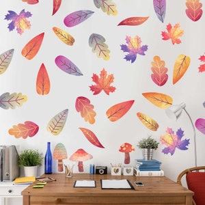 May include: A white wall with watercolor leaf decals in shades of pink, orange, red, and purple. A desk with a wooden top and a white base is in front of the wall. The desk has a lamp, a vase with flowers, and a few other items on it.
