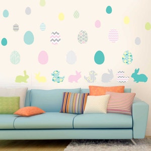 May include: A light blue couch with colorful patterned pillows in front of a white wall with pastel Easter egg decals. The decals feature various patterns, including stripes, polka dots, and chevron.