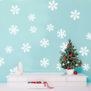 May include: White snowflake wall decals on a light blue wall with a small artificial Christmas tree decorated with red and blue ornaments. The tree is in front of a white dresser with drawers.