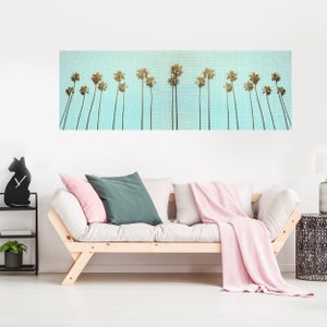 May include: A panoramic print of palm trees against a blue sky with a white grid pattern. The print is hanging above a light wood couch with pink and green throw pillows and a pink throw blanket.