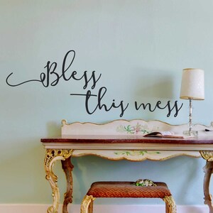 May include: A black vinyl wall decal with the text "Bless this mess" in a cursive font. The decal is placed on a light blue wall above a white desk with a floral design.