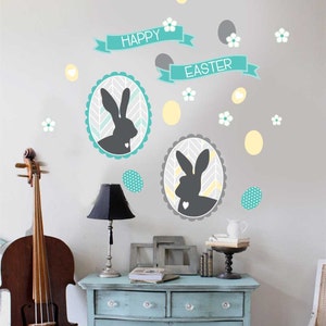 May include: Wall decals with a Happy Easter theme. The design includes two oval frames with gray bunny silhouettes, teal banners, and decorative eggs and flowers. The color palette features gray, teal, yellow, and white.