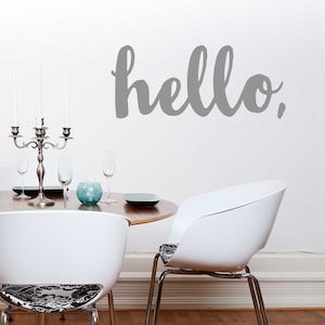 May include: A gray vinyl wall decal with the word "hello" in a handwritten style. The decal is on a white wall in a dining room with a table and chairs.
