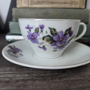 May include: White teacup and saucer set with a floral design featuring purple flowers and green leaves. The teacup and saucer have a gold trim. The teacup is sitting on a wooden surface.