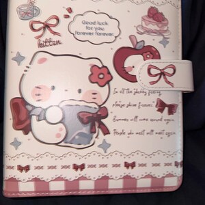 Puede incluir: Una agenda blanca y rosa con un diseño de gatito de dibujos animados. La agenda presenta ilustraciones de una taza de té, un pastel y una manzana. El texto incluye "Good luck for you forever forever". Un cierre en forma de lazo asegura la agenda.