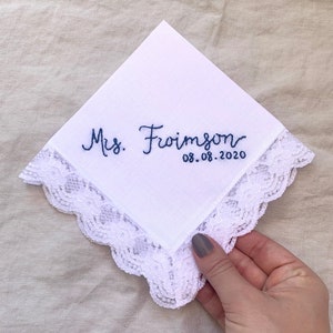 Image shows a white handkerchief with a detailed edge, folded into quarters. The text shows "Mrs. Froimson" along with the date, embroidered in blue thread.