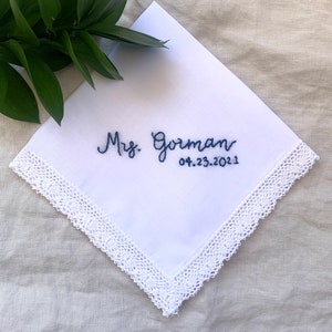 Image shows a white handkerchief with a detailed edge, folded into quarters. The text shows "Mrs. Gorman" along with the date, embroidered in blue thread.