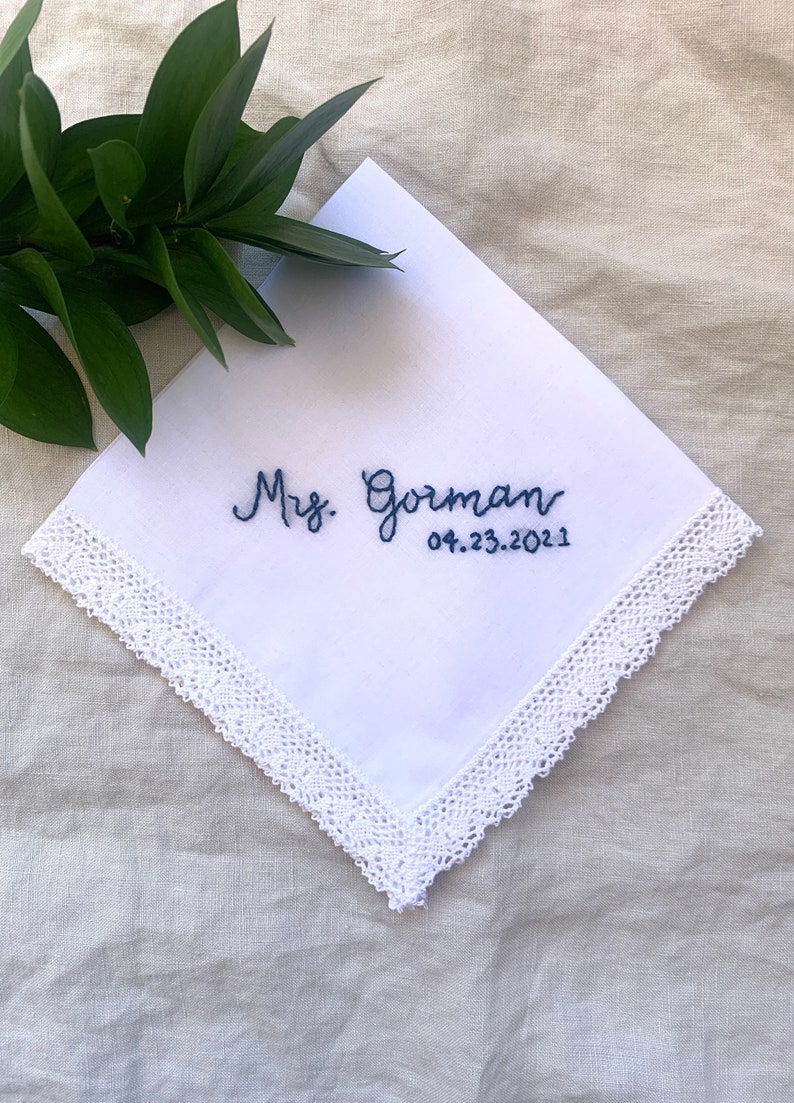 Image shows a white handkerchief with a detailed edge, folded into quarters. The text shows "Mrs. Gorman" along with the date, embroidered in blue thread.