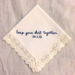 Image shows a white handkerchief with a detailed edge, folded into quarters. The text reads keep your shit together in blue embroidery thread, with the date underneath.